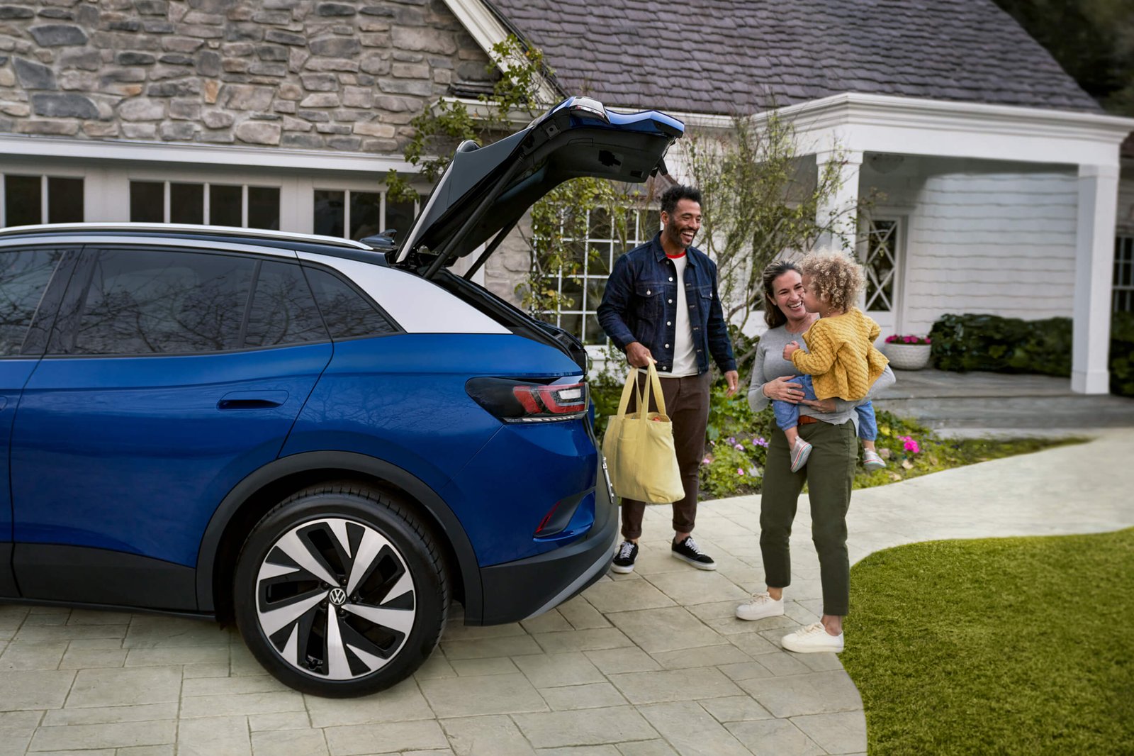 A young family loads a bag into the cargo area of an ID.4, shown in Dusk Blue Metallic parked in a residential driveway.