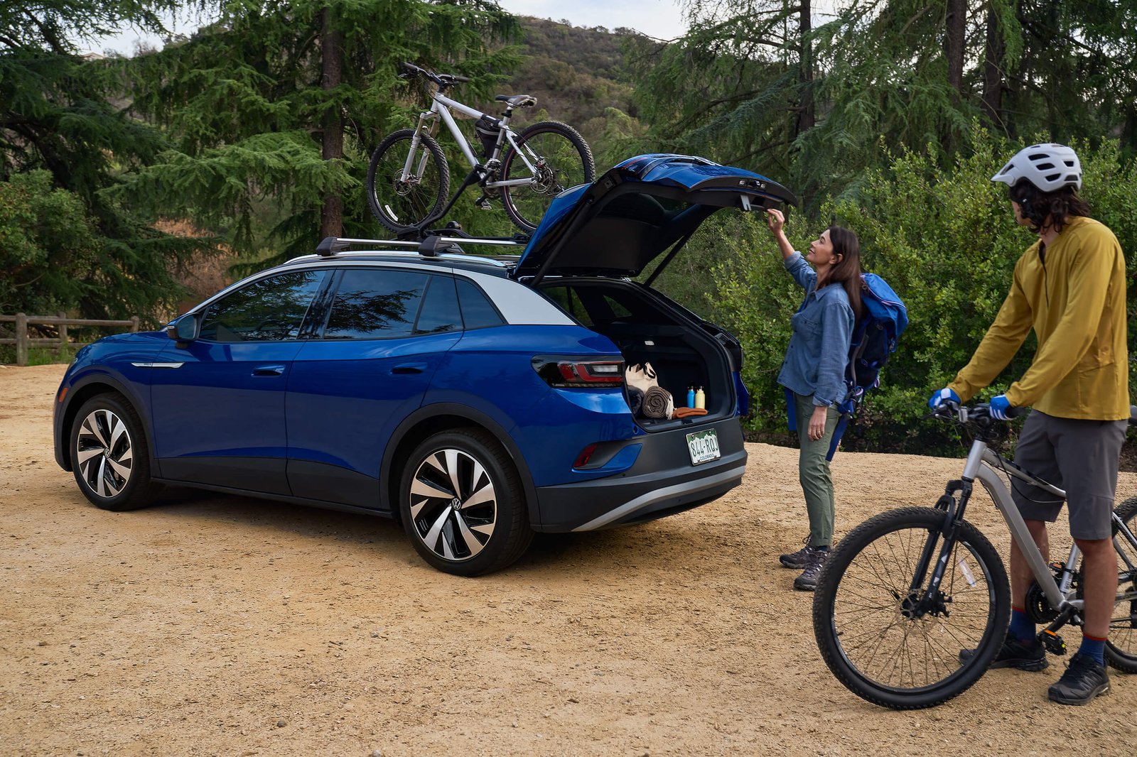 A man and woman load bicycles onto an ID.4, shown in Dusk Blue Metallic with accessory bicycle racks and parked in a natural setting.