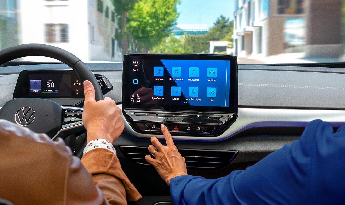 A close-up shot of an ID.4 SUVdashboard over the shoulders of thedriver and passenger, focusing on thecenter display as thepassenger reaches out to the touchscreen.